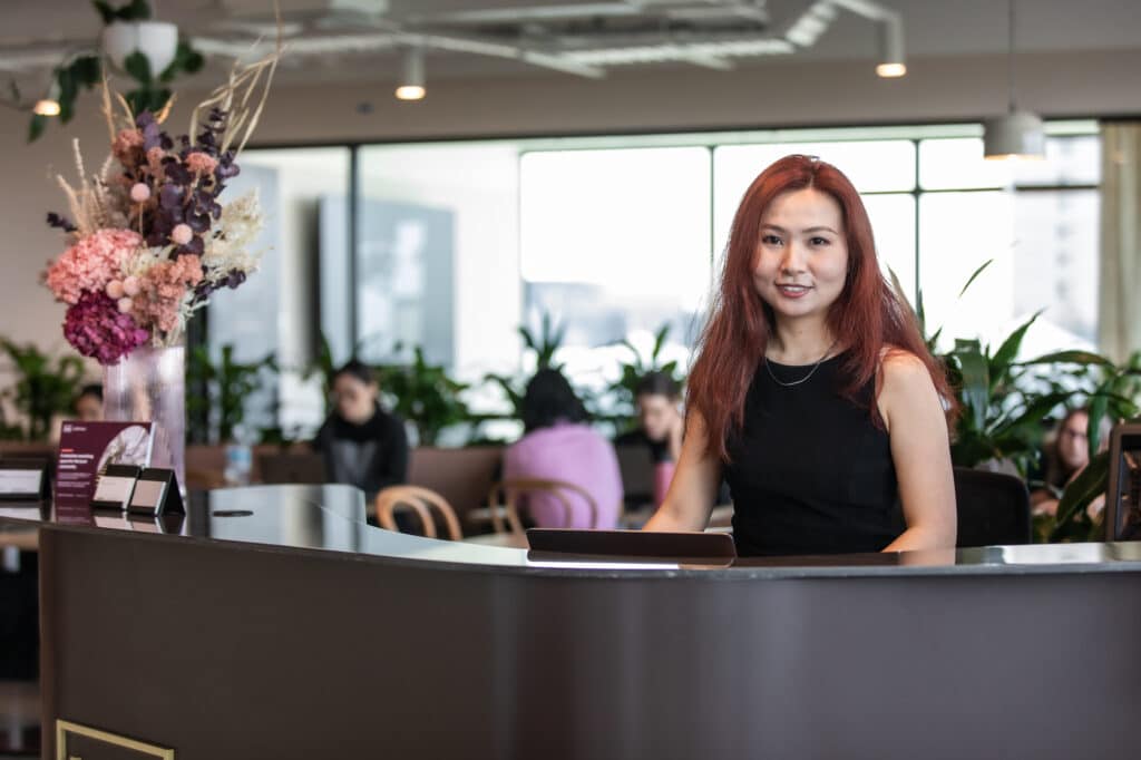 Young Asian woman smiling behind the welcome desk at Hub Local Box Hill, ready to assist visitors.