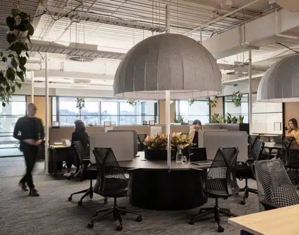 A shared office space featuring a range of tables. There is a woman sitting at a desk on her laptop.