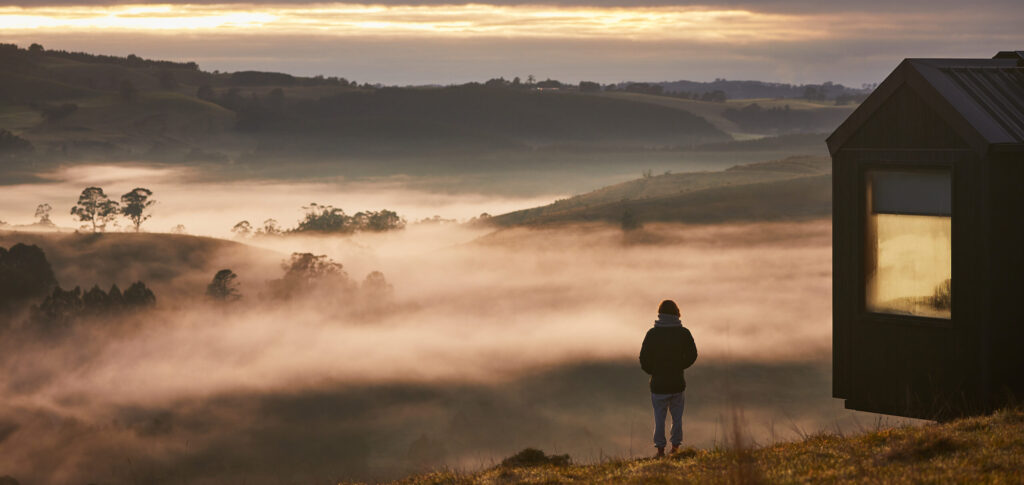 frosty, smog view of land with a man standing watching and an unyokd cabin overlooking